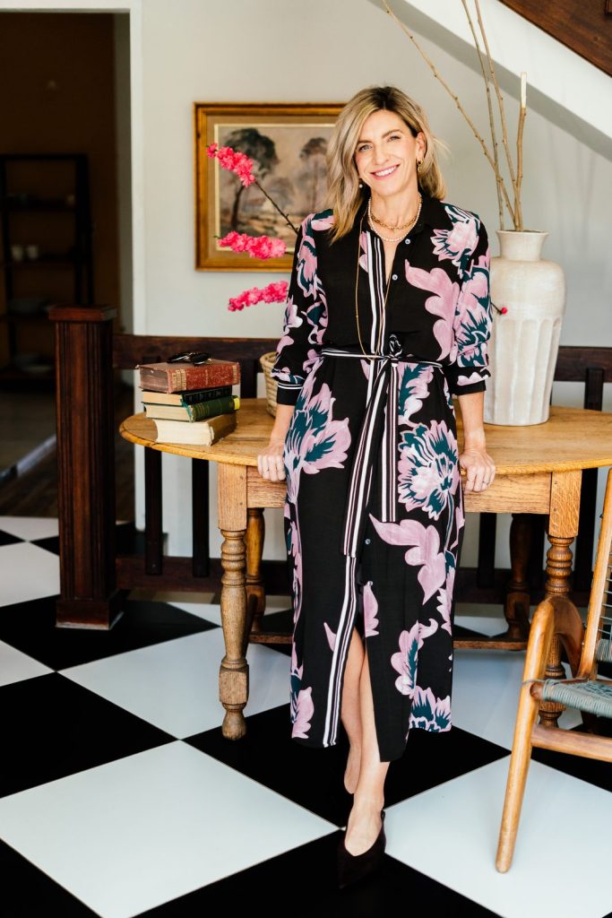 Jeanine in a black floral dress, standing by a table on a chequered floor