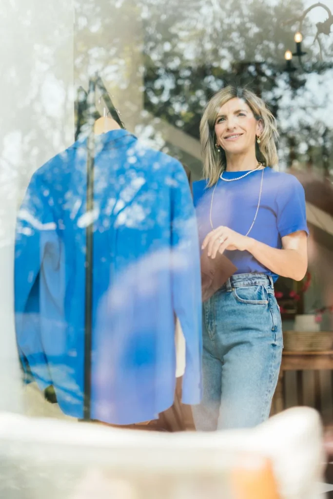 Jeanine seen through a window in a cobalt tee and jeans, beside a blue shirt on a hanger